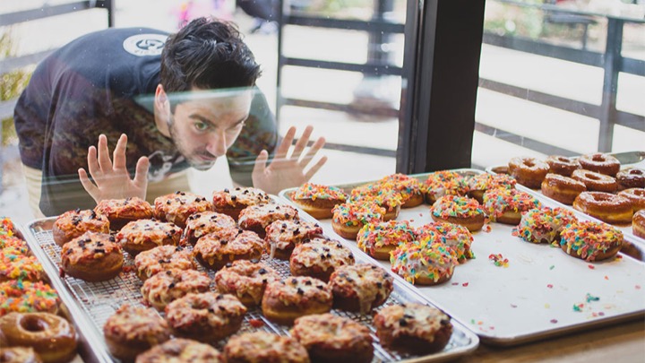 A white male with brown hair in a black t-shirt, outside of a pastry shop, with his hands against the glass looking at a tray of donuts